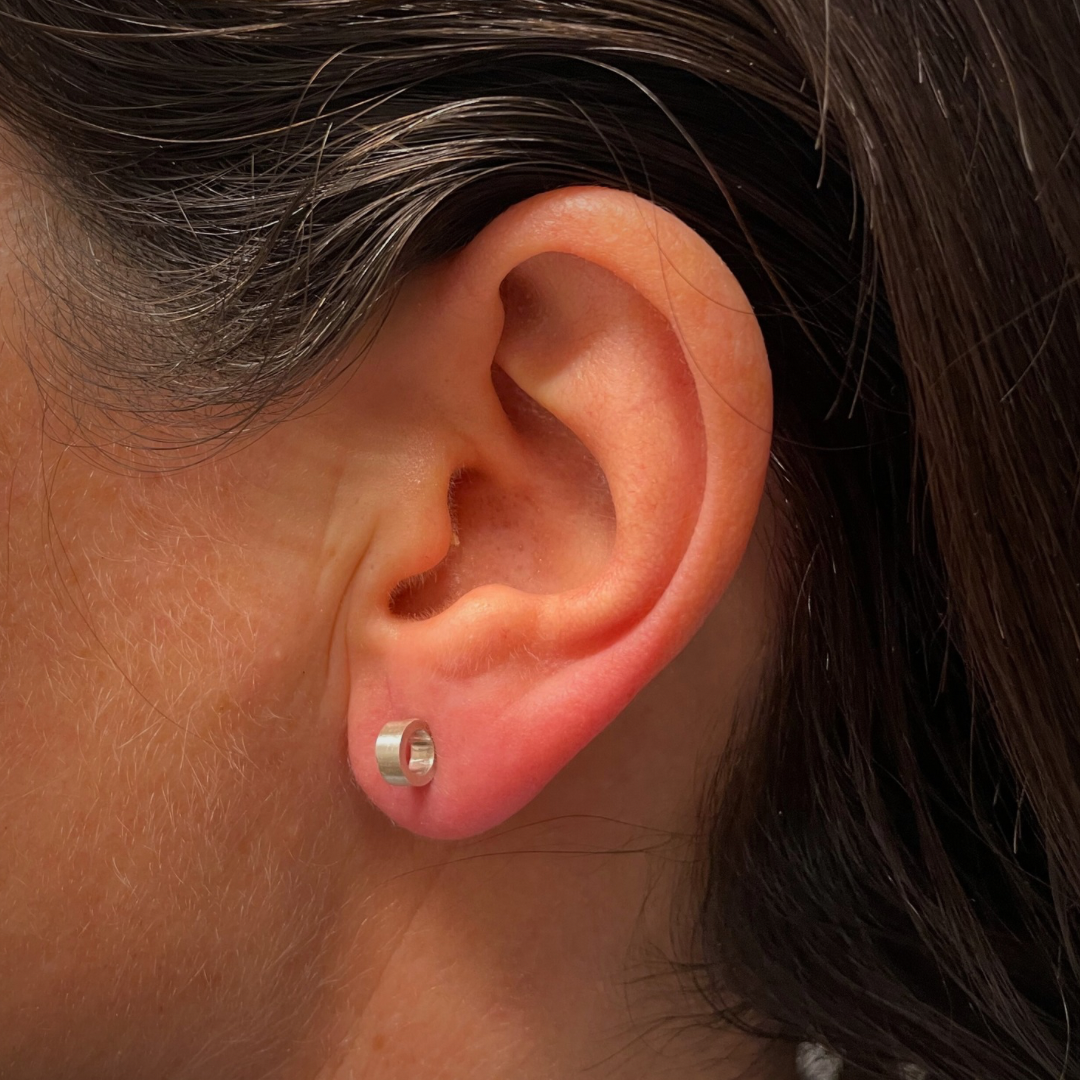 Close up of a woman’s ear who is wearing a sterling silver earring with a circular shape that has a diameter of 6 mm and which is placed perpendicular to the ear.